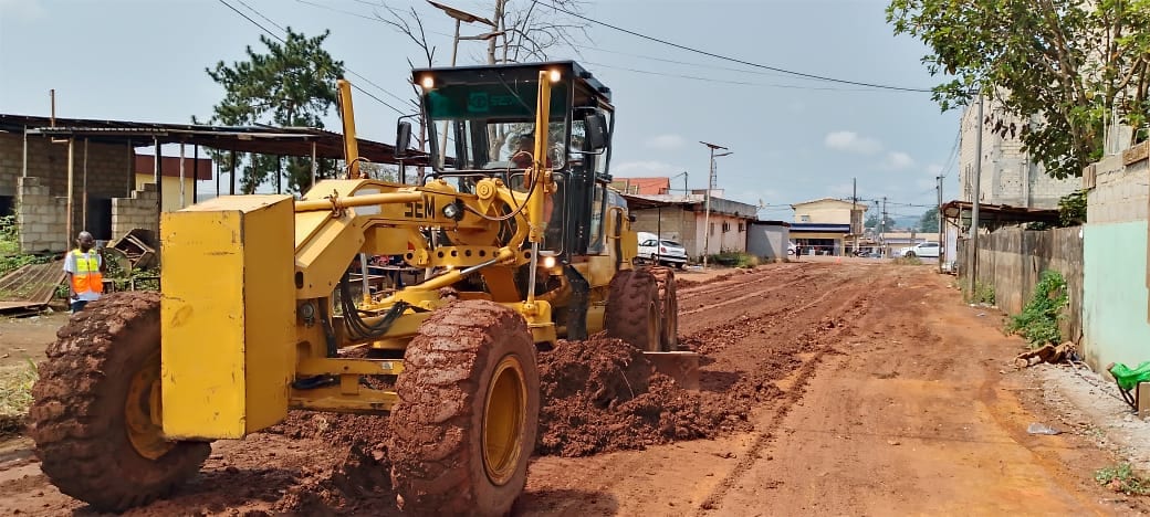 Engin de chantier reprofilant une voie secondaire non bitumée.
