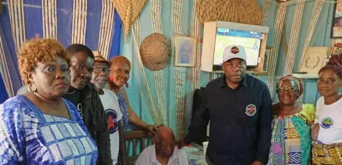 Un homme centenaire assis chez lui reçoit la visite de membres d'une association gabonaise venus lui apporter des produits de première nécessité.
